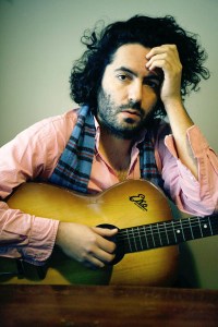 daniel-bejar-destroyer-in-pink-shirt-and-plaid-scarf-holding-guitar-against-green-background-1 Daniel Bejar Destroyer in pink shirt and plaid scarf holding guitar against green background