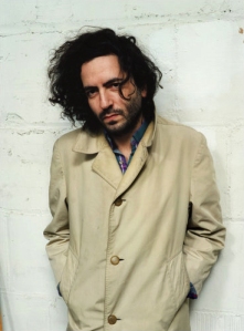 Daniel Bejar Destroyer leaning against white wall in a trenchcoat